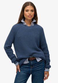 Blauwe gebreide trui met ribbeltextuur, ronde halslijn en ingezakte schouders, gedragen over een lichtblauw gestreept shirt, gecombineerd met jeans.