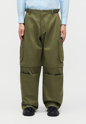 Vyras, dėvintis alyvuogių žalios spalvos krovinių kelnes su daug kišenių, juodus sportinius batelius ir šviesiai mėlyną marškinius su sagomis, stovintis prieš tuščią baltą foną.