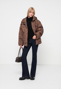 Hnědá pufovaná bunda s leopardím vzorem a kapucí, černý svetr a rozšířené tmavé džíny. Drží malou černou kabelku a má na sobě černé boty.
