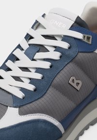 Γκρι και μπλε αθλητικό παπούτσι με λευκούς κορδόνια και σόλα, με μεταλλικό λογότυπο "B" στην πλευρά και λευκή ετικέτα στη γλώσσα.