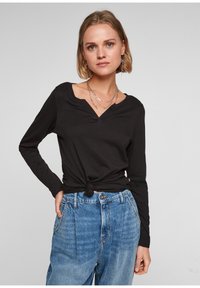 Schwarzes Langarmshirt mit V-Ausschnitt und geknoteter Front, kombiniert mit hellblauen hochgeschnittenen Jeans. Weicher Stoff, taillierte Passform.