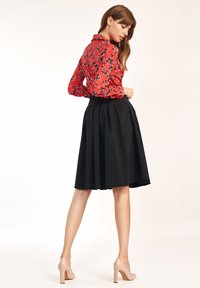 Kobieta w czerwonej kwiecistej bluzce, czarnej plisowanej spódnicy midi i beżowych szpilkach, pozująca na tle gładkiej białej ściany.