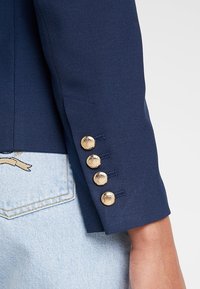 Navy blue -takki, jossa on teksturoitu kangas. Varustettu neljällä kultaisella napilla hihassa ja muotolennetulla istuvuudella. Hihan päättyy ranteen yläpuolelle.