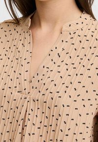 Blouse beige avec plis verticaux, ornée de motifs de nœuds noirs. Tissu léger, encolure en V et manches courtes.