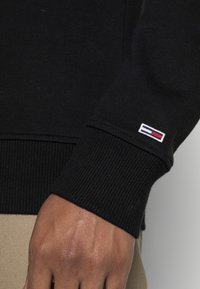Manica di maglione nero lavorato a maglia con polsino a costine e piccola toppa logo in rosso, bianco e blu sul lato sinistro. Primo piano della mano e della trama del tessuto.