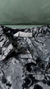 Črno-bela bluzica s prišitim ovratnikom in gumbnim zapiranjem. Tkanina prikazuje marmorni vzorec z gladko teksturo. Oznaka za obešanje je vidna.
