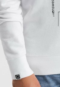 Sudadera blanca con puños y dobladillo acanalados, que presenta un detalle de logo impreso. Textura suave, corte relajado y diseño casual.