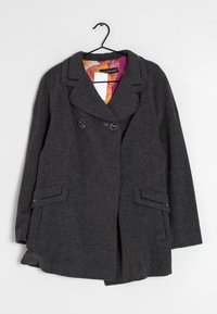 Cappotto di lana grigio con bottoni doppi, ampio colletto, tasche laterali e una fodera interna con un motivo colorato. Appeso a un gancio.