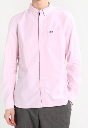 Mand iført en lyserød langærmet skjorte med knapper og en lille grøn krokodille-logo på brystlommen, kombineret med grå bukser.