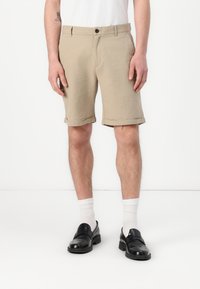 Beige Leinenshorts mit umgeschlagenen Säumen, einem Frontknopfverschluss, Seitentaschen und einem lockeren Schnitt, kombiniert mit schwarzen Slippern und weißen Socken.