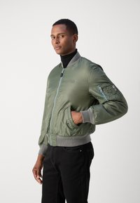 Giubbotto bomber verde in nylon con chiusura a zip, polsini a coste grigi e due tasche laterali; presenta una toppa sulla manica.