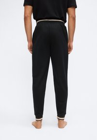 Άτομο που στέκεται ξυπόλυτο με την πλάτη στραμμένη, φοράει μαύρο παντελόνι τζόγκερ με ρίγες στα μανσέτα και ταιριαστό μαύρο πουκάμισο.