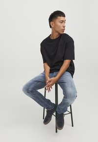 T-shirt nera in cotone, vestibilità rilassata, abbinata a jeans blu sbiaditi. Seduto su uno sgabello di metallo nero, indossa sneakers in suede scure.