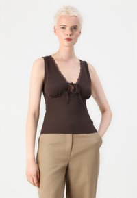 Top marrone senza maniche con scollatura a V, bordo in pizzo e dettaglio con fiocco sul seno. Tessuto texturizzato, stile aderente, abbinato a pantaloni color taupe.
