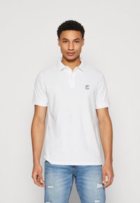 Hvid polo skjorte med korte ærmer, der har en krave og et lille logo på venstre bryst. Bæres sammen med lyseblå slidte jeans.