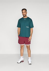 Türkiz, rövid ujjú póló bal oldalon logóval, bordó sort fehér díszítéssel, és fekete akcentusos fehér edzőcipő.