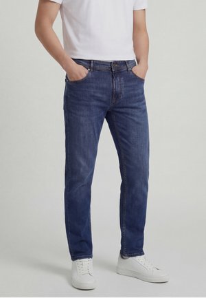 Man wearing straight-fit blue jeans, white t-shirt, and white sneakers, standing with hands in pockets against neutral background.