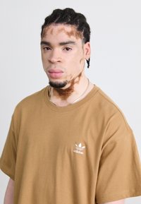 Tricou din bumbac maro cu mâneci scurte, având un logo alb Adidas în zona stângă a pieptului. Textură netedă, cu o croială lejeră.