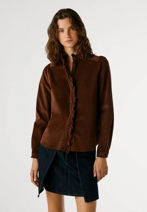 Camicia di velluto a coste marrone con una placca arricciata, chiusura a bottoni e borchie sulle spalle, abbinata a una gonna di jeans scura con uno spacco.