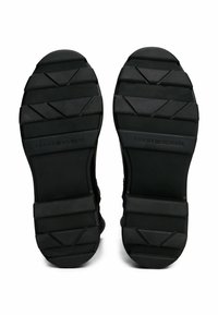 Zwarte rubberen schoenenzolen met een gestructureerd grippatroon; bevat hexagonale en driehoekige vormen, en het reliëf van het merk "TOMMY HILFIGER."