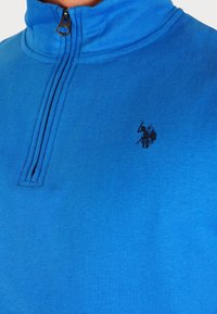 Blauwe fleece pullover met een kwart-ritssluiting en hoge kraag, voorzien van een tonale logo op de borst. Glad textuur en casual ontwerp.