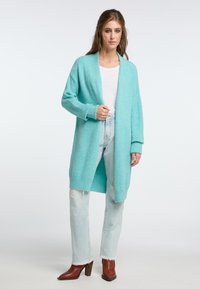 En ljusblå, öppen cardigan i mjukt material, med långa ärmar och en avslappnad passform. Tillsammans med ljusa slitna jeans och bruna stövlar.