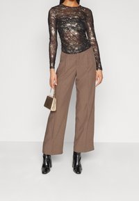 Schwarzes, glitzerndes Langarmoberteil mit gerafftem Design, kombiniert mit weiten, braunen Hosen. Accessoires sind eine kleine goldene Handtasche und schwarze Stiefel.