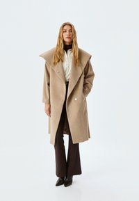 Abrigo de mezcla de lana beige con un gran cuello tipo chal, frente de doble botonadura y botones en tono plateado, combinado con pantalones oscuros y botas.