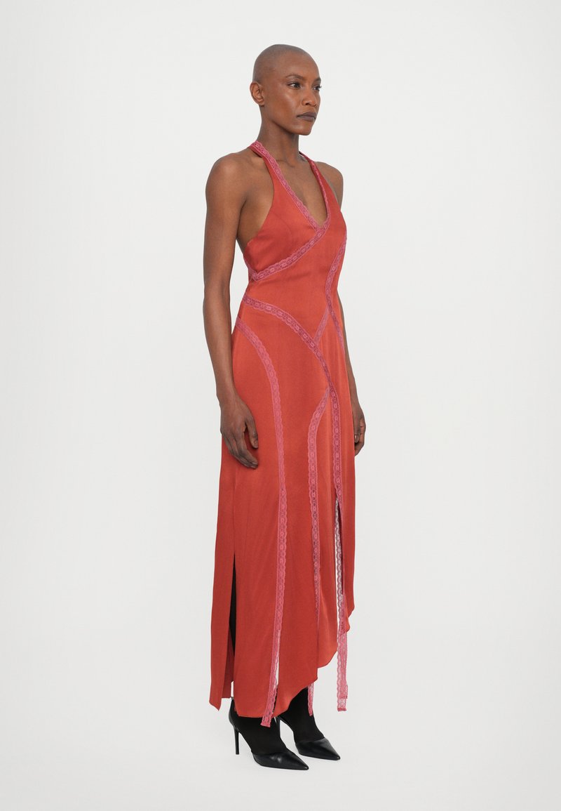 Lungo abito rosso con accenti in pizzo, scollatura all'halter e spacchi laterali. Tessuto morbido, silhouette aderente, abbinato a scarpe nere con tacco alto.