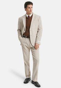 Mann in hellbeigem Anzug mit Jackett und Hose, braunem Pullover über weißem Hemd mit roter Krawatte, schwarzen Schuhen, stehend mit einer Hand in der Tasche.