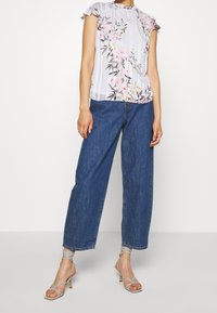 Blus i ljusgrått med blommönster och volangärmar, tillsammans med vida blå denimjeans. Accentuerad med grå sandaler med remmar.