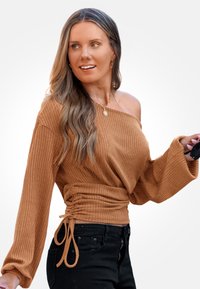 Brązowa, prążkowana bluzka bez ramiączek z długimi rękawami, ze strefą w talii, w połączeniu z czarnymi spodniami i trzymająca okulary przeciwsłoneczne.