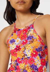 Top de tirantes con estampado floral que presenta un patrón vibrante de flores rojas, moradas, amarillas y blancas sobre un fondo lila. Tirantes finos; diseño ajustado.