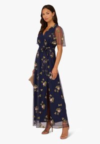Marineblaues Blumenwickelkleid mit durchsichtigen Ärmeln und einem gebundenen Taillengürtel. Es verfügt über rosa und grüne Stickereien sowie einen Schlitz bis zum Oberschenkel. Aus glattem, leichtem Stoff.