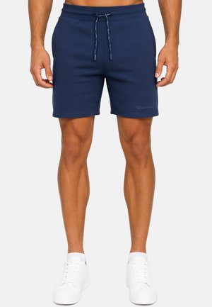 Modello maschile che indossa pantaloncini blu navy con coulisse e sneakers bianche, in piedi davanti a uno sfondo chiaro e uniforme.