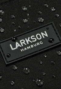 Tessuto nero con gocce d'acqua e un'etichetta rettangolare nera con "LARKSON HAMBURG" in lettere bianche in rilievo. Superficie texturizzata.