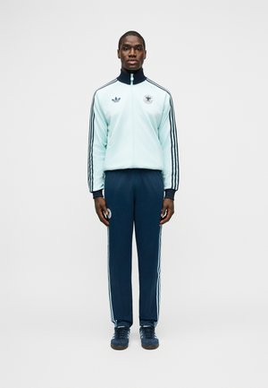 Mees seisab, kandes helehalli ja tumesinisega spordiriietust, millel on Adidas logo ja triibud, suunaga otse eest, üksluisel taustal.