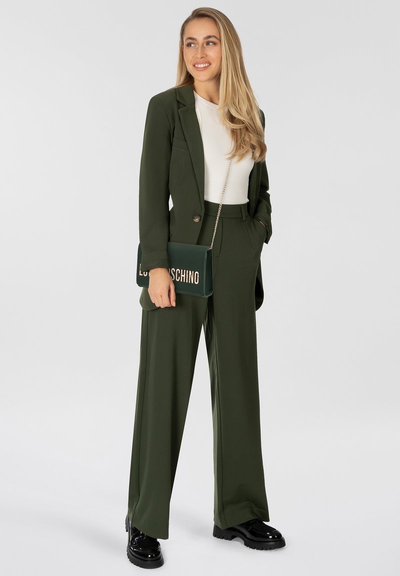 Completo verde oliva con giacca aderente, pantaloni a gamba larga e top bianco. Tiene una borsa verde con scritte dorate. Scarpe con piattaforma nere.