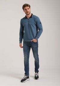 Hele sinine pehme pool tõmblukuga pullover, tekstuuriga rinnaosa ja must kaelus. Kanda siniste teksade ja mustade tossudega.