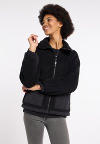Zwarte sherpa-jack met gevoerde stofaccenten, ritsluiting, hoge kraag en elastische manchetten. Heeft een zachte textuur en een relaxte pasvorm.