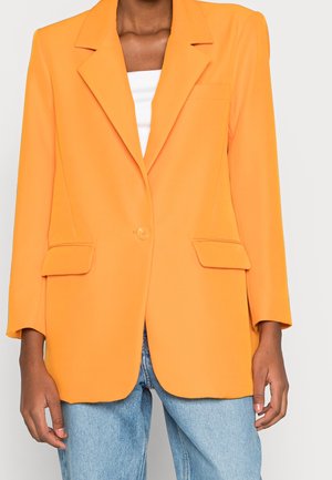 Personne portant un blazer orange vif à un seul bouton sur un top blanc, associé à un jean bleu clair, montré des épaules aux hanches.