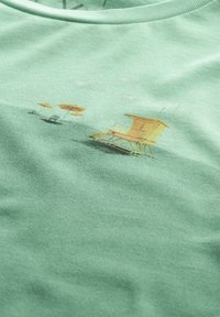 Groene katoenen T-shirt met een grafische print van gele strandreddingtoren en parasols op een strandscène, met een lichte textuur als achtergrond.