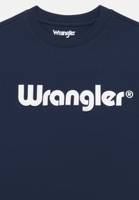 T-paita, jossa on laivastonsininen väri ja joka on valmistettu puuvillasta. Etupuolella on suuri valkoinen "Wrangler"-logo. Pyöreä kaula-aukko ja lyhyet hihat.
