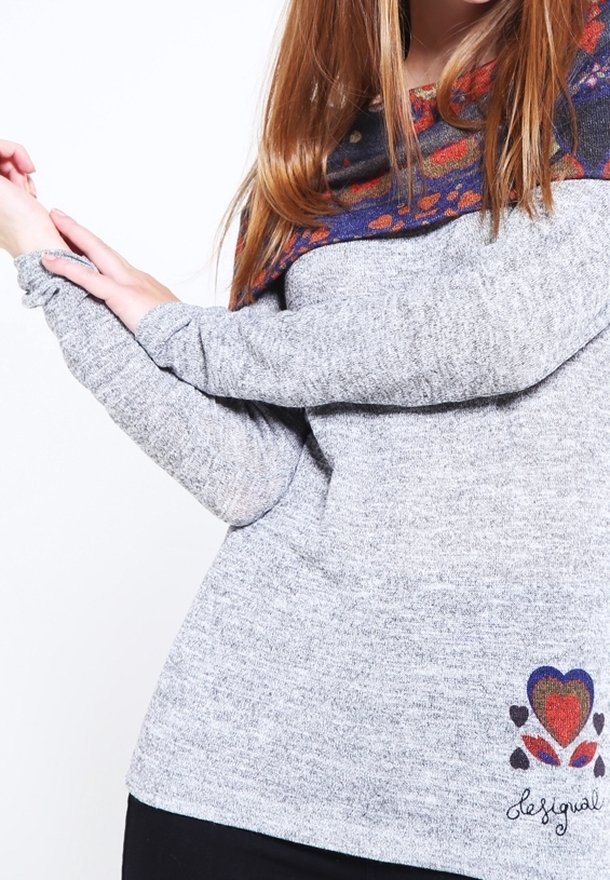 Personne portant un pull gris avec un col à motifs colorés et un motif en forme de cœur près de l'ourlet, étirant son bras gauche à travers le corps.
