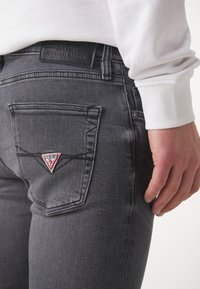 Jeans de mezclilla gris con una vista en primer plano del bolsillo trasero y un parche de logo triangular. La tela parece suave con un aspecto ligeramente deslavado.