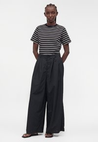 Femme debout, mains derrière le dos, portant un haut à manches courtes rayé noir et blanc, un pantalon noir à jambes larges et des sandales noires.