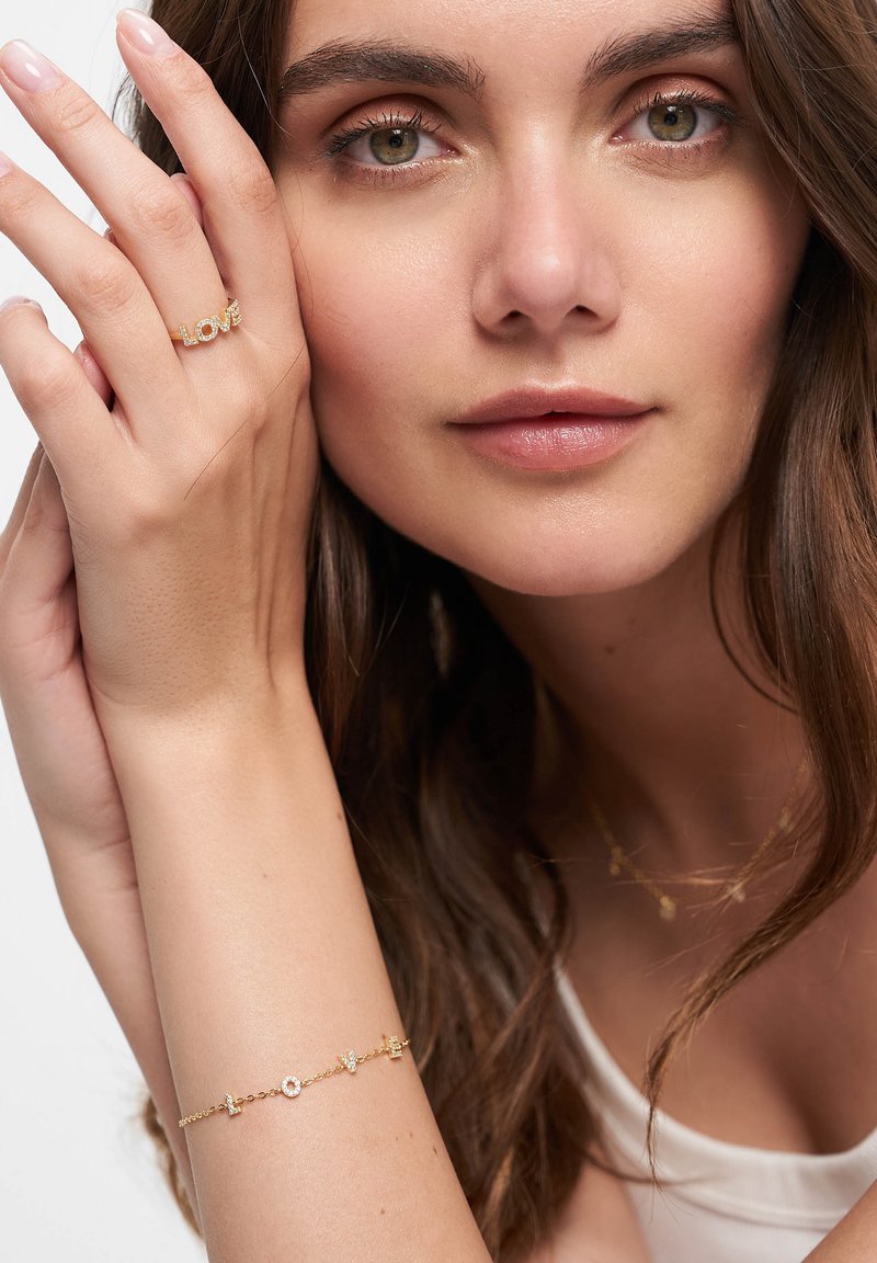 Guldjewelry inkluderar en ring med texten "LOVE" och ett delikat armband med små detaljer. Modellen har långt vågigt hår och en neutral klädsel.