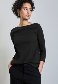 Schwarzes gestreiftes Langarmoberteil mit einem Bootsausschnitt-Design. Weicher Stoff, figurbetonte Silhouette und leichtes Drapieren am Saum. Kombiniert mit schwarzen Hosen.