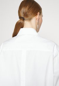 Camicia bianca con chiusura a bottoni, ricamata con "KARL LAGERFELD" sul retro, texture liscia, vestibilità classica, con dettagli plissettati.