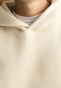 Felpa beige con una texture morbida, caratterizzata da un cappuccio regolabile e indossata sotto una camicia bianca. Primo piano della zona del collo e dei dettagli dell'indumento.